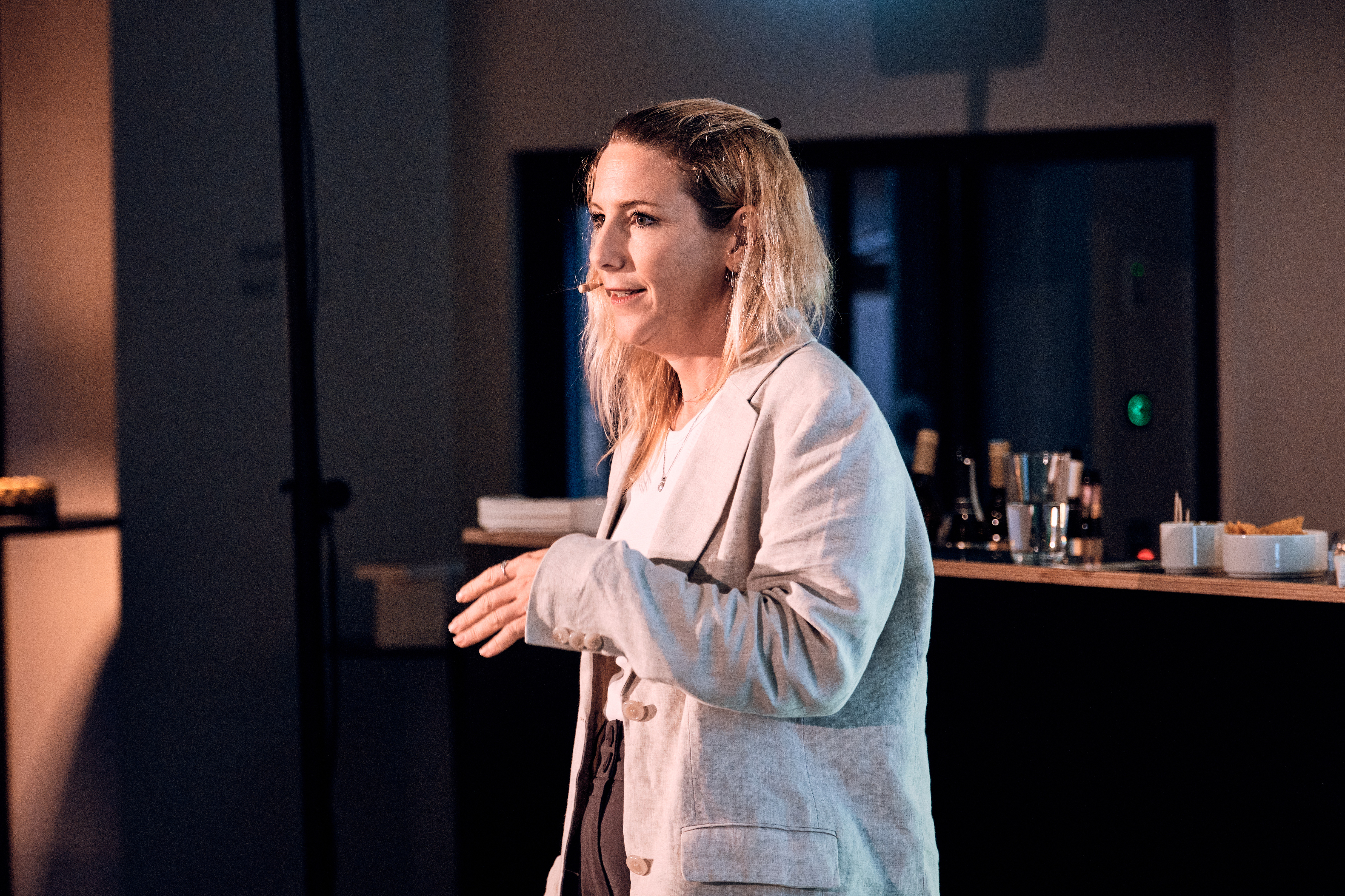 Eine Frau in einem hellen Blazer spricht in ein Headset-Mikrofon und steht in einem Innenraum neben einem Bartresen.